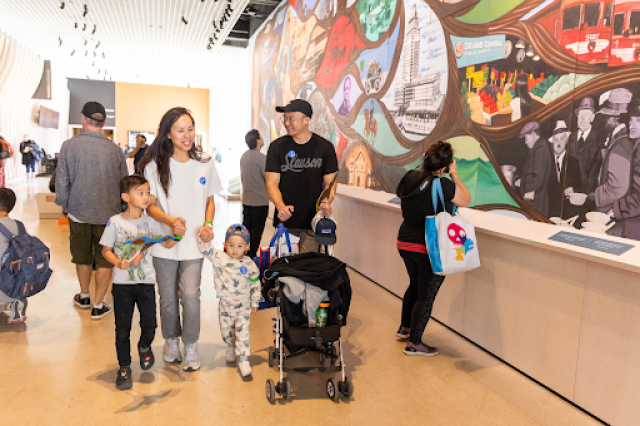 Family of man pushing stroller and woman holding toddler and older child's hands walking past Carasco mural in NHM Commons