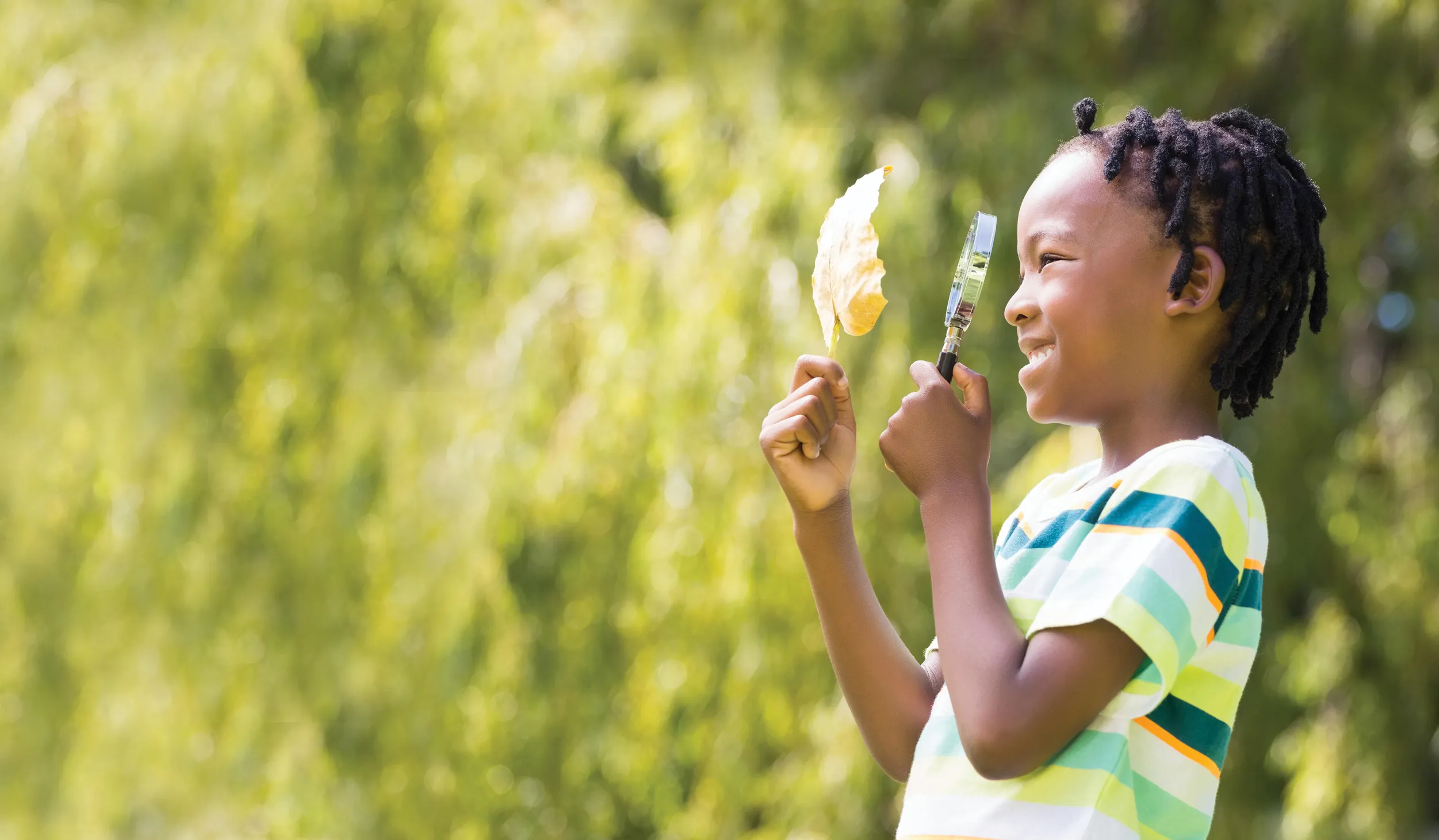 Side view of a child looking at leaf through a magnifying glass, against a blurry leafy green background