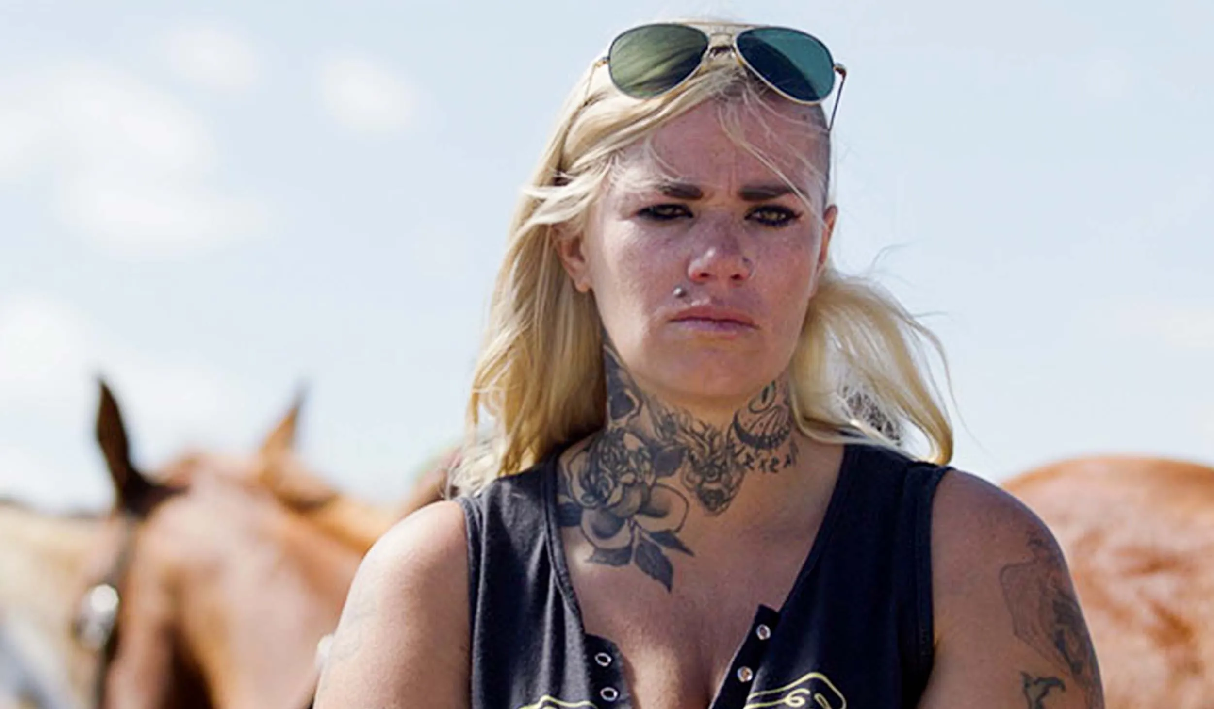 Head and shoulders of blonde haired woman with sunglasses on her head, wearing a black tank top, with a the head of the horse facing away from the camera in the background