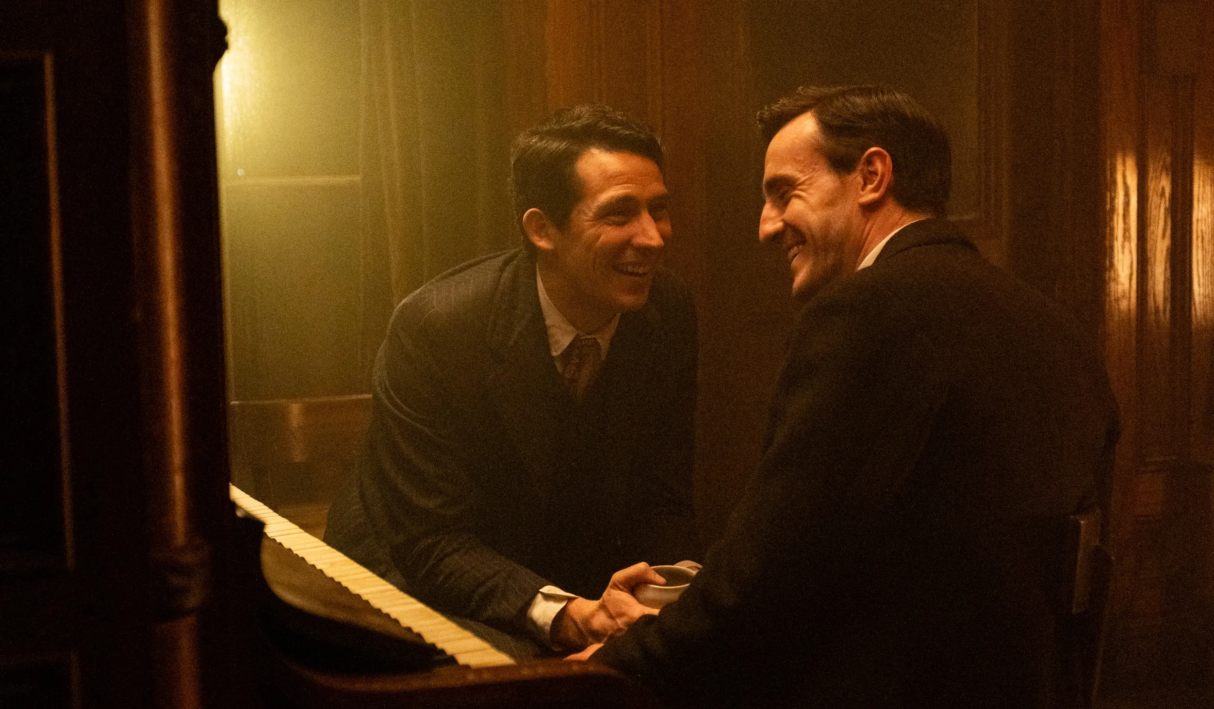 Two men in suits sitting by a piano, facing each other
