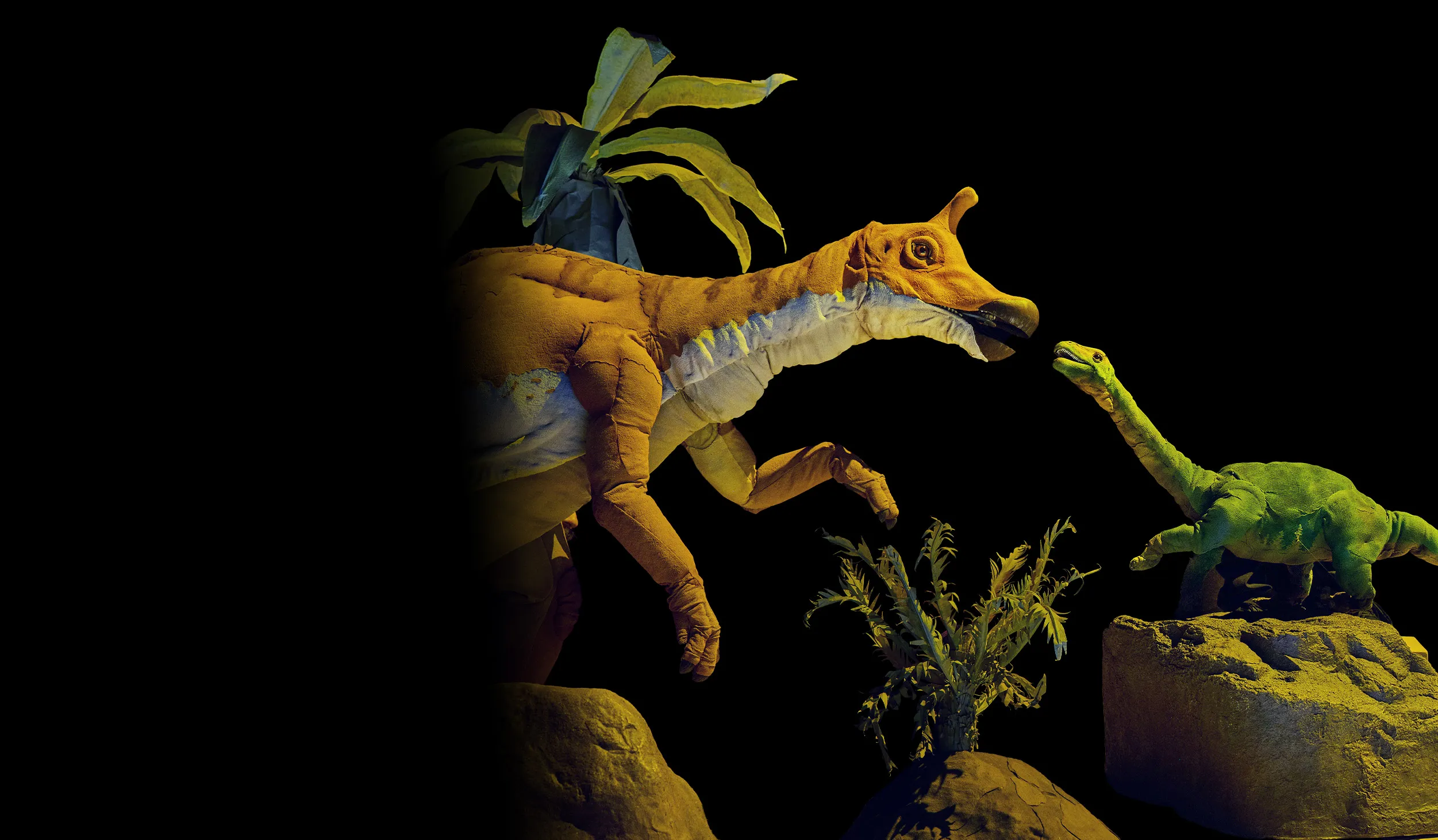 Hadrosaur orange and white puppet facing a green miniature long-necked dinosaur puppet