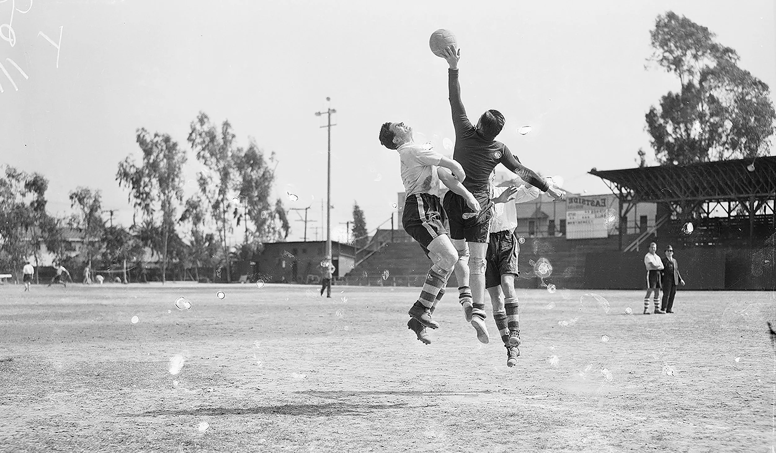 Black and white photo of three soccer players jumping in the air while one player reaches for the ball with his hand