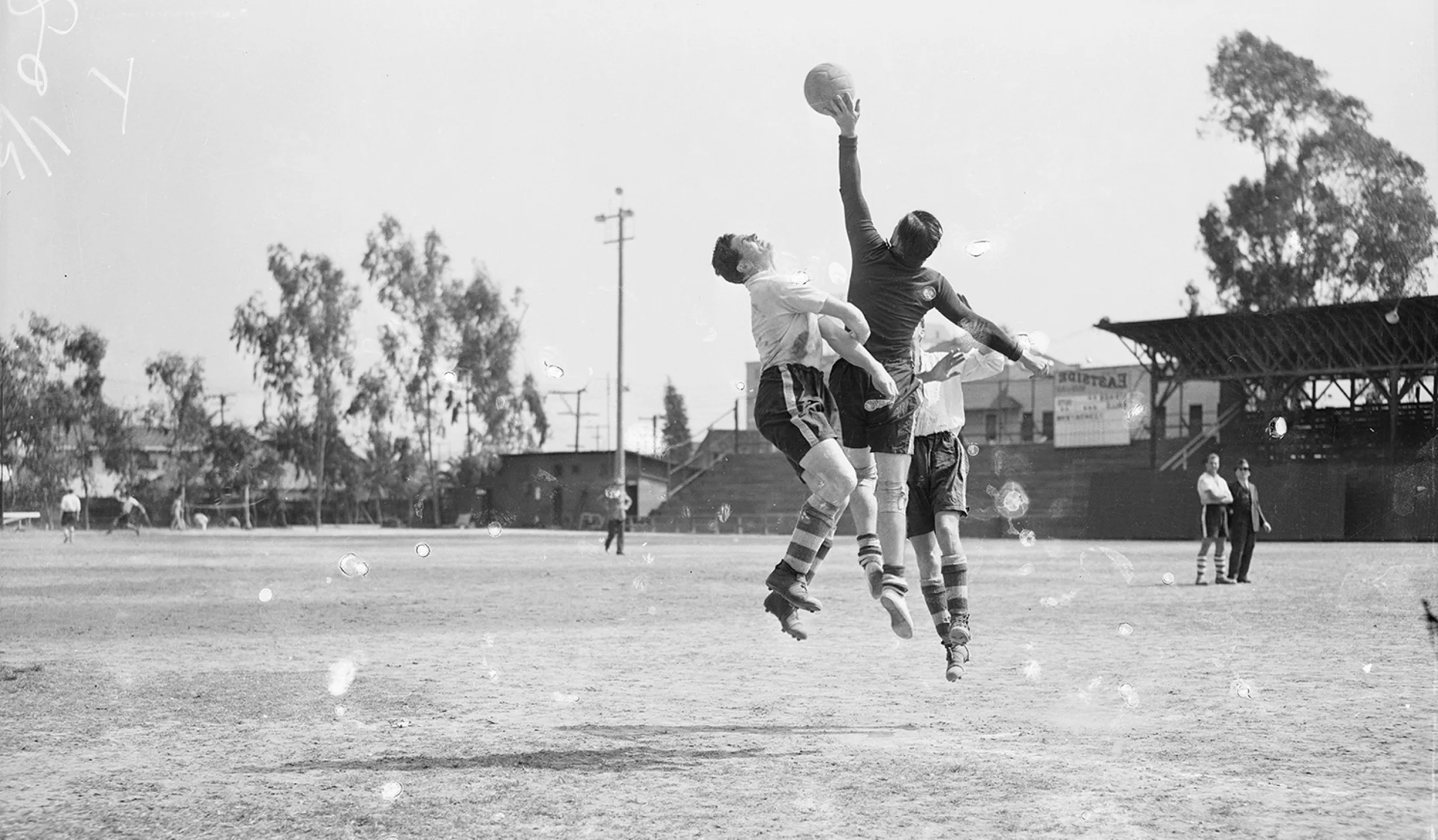 Black and white photo of three soccer players jumping in the air while one player reaches for the ball with his hand