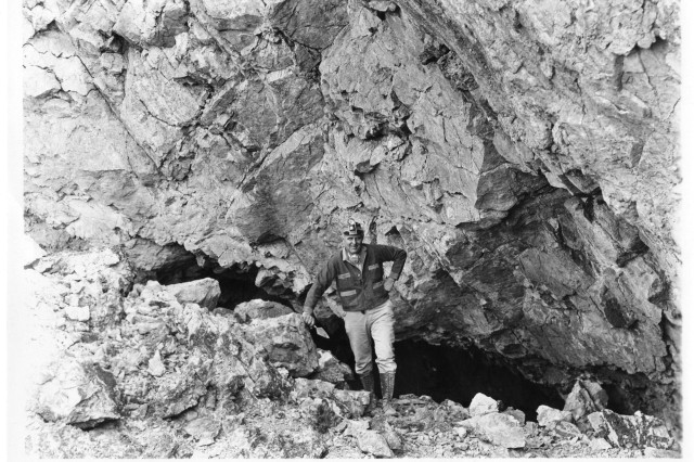 Man holding trowel stood outside the mountainous entrance to a cave.