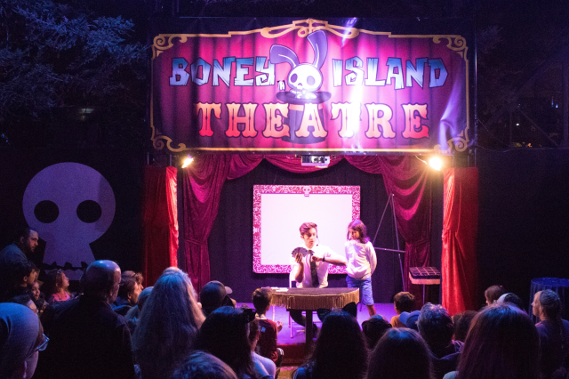 Audience members seen from behind, facing a stage with two performers with a sign above that reads Boney Island Theatre in capita letters with an illustration of a rabbit coming out of a top hat