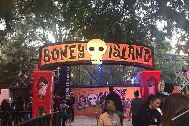 Visitors walking through a gated entryway with a banner with text in capital letters and an illustrated skull between Boney and Island on an orange background.