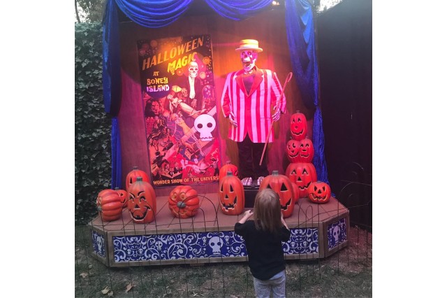A child seen from behind, facing a stage with pumpkins and a replica skeleton wearing a striped jacket and a hat