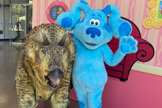 Person wearing a blue dog costume with its arm around a human-sized dinosaur puppet