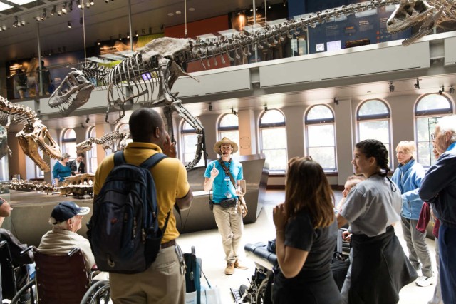 Group of visitors, some in wheelchairs, facing a Museum Educator with dinosaur skeletons in the background