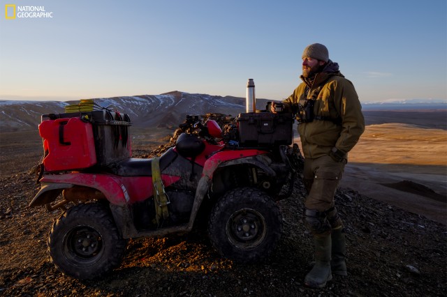Photographer with a camera around their neck, holding the top of a thermos and standing by an off-road vehicle with a thermos on top