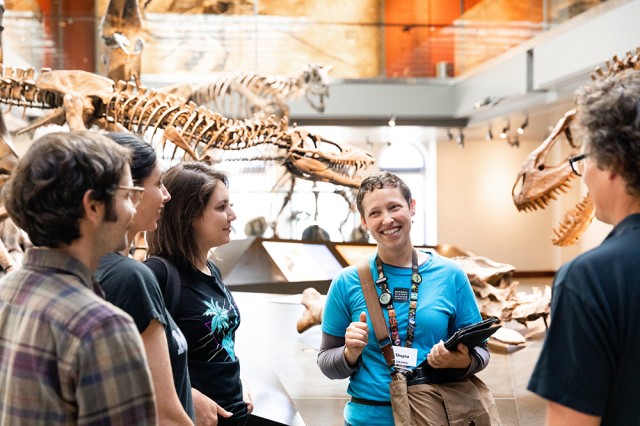 Visitors facing a Museum Educator with dinosaur skeletons in the background