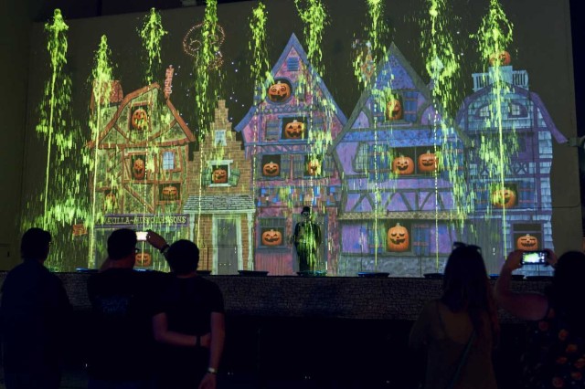 Visitors watching a cut-out of houses with pumpkins in the window and green water spraying upwards in front of it 