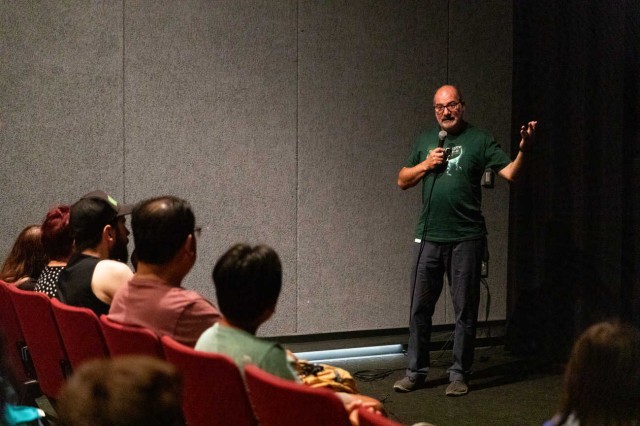 Luis Chiappe talking with a microphone, facing guests in red seats
