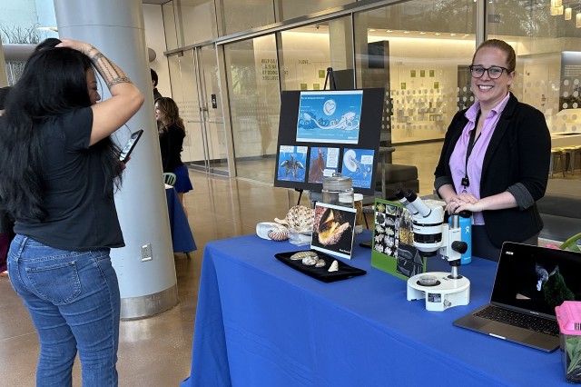 Student looking at a table display of ocean specimens and signage, with an NHM staff member smiling behind the table