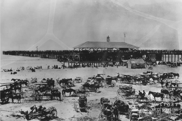 Antique photo of dozens of wagons with people and horses parked on the beach next to a pier