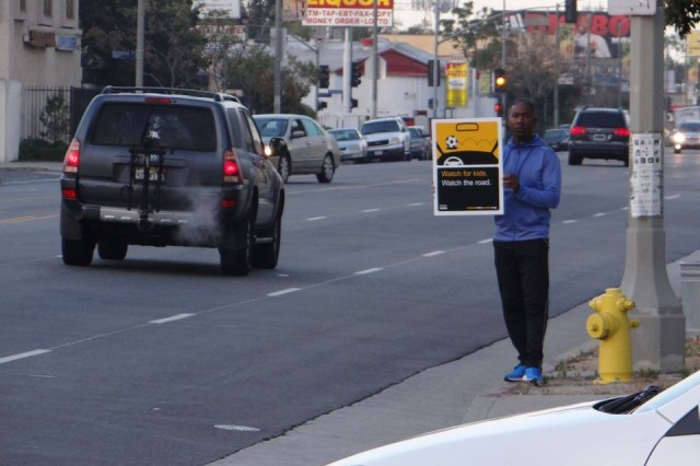 Man of color in a blue sweatshirt and black sweatpants standing on a street corner holding a sign that says &quot;watch for kids. Watch the road.&quot; as cars drive past.