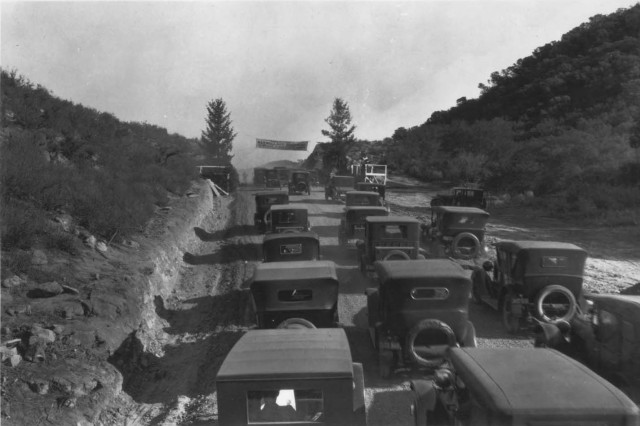 Looking from behind at a three-lane dirt road filled with Model T cars and grassy hills on either side of the road.