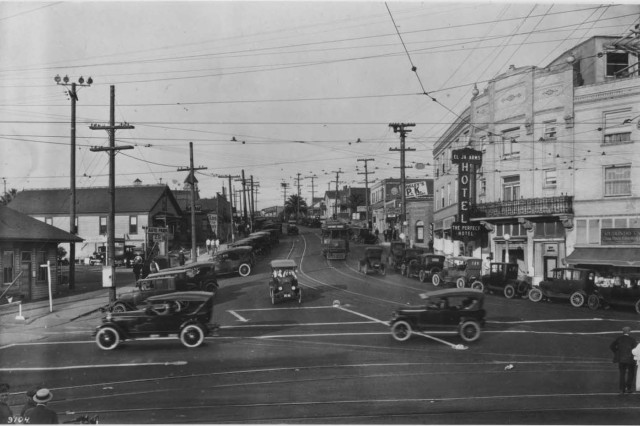 Black and white photograph of a T-shaped intersection filled with Model T cars. Small houses line the left side of the street and three-story commercial buildings are on the right. Telephone wires crisscross above the street. 