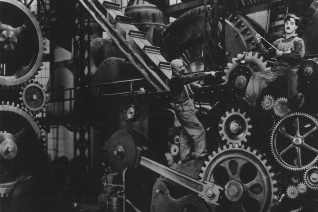 Black and white photograph of Charlie Chaplin sitting on a giant machine make of cogs and gears while another man climbs up the wheels pointing at him.