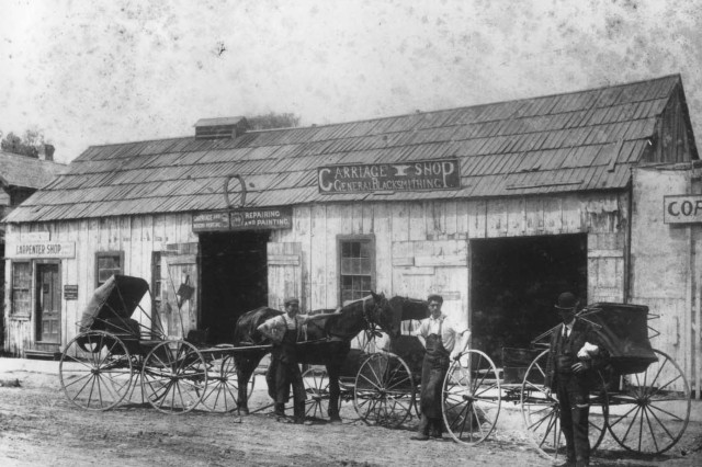 Antique photo of three men and one horse standing alongside three carriages in front of a rustic wooden building under a sign that reads &quot;Carriage Shop General Blacksmithing&quot;