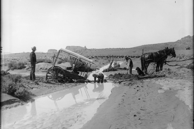 Antique photograph of covered wagon stuck in a muddy creek. Two horses stand on the opposite shore while three men try to dig the front of the wagon out of the mud.