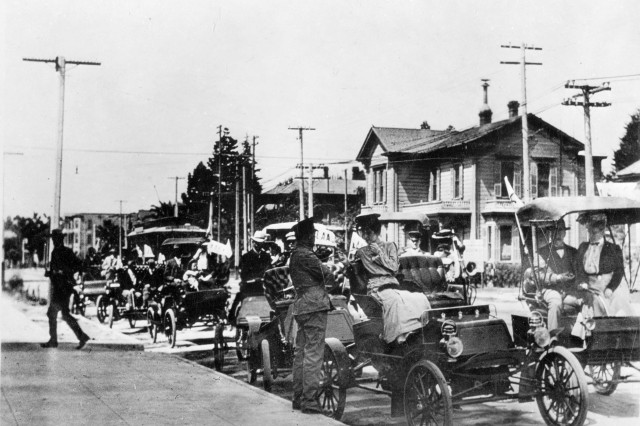 Antique photo of eight motorized carriages parked along the edge of the street with men and women standing around or sitting in the car in late 19th century dress. A large wooden two-story house, trolly car, and telephone poles are visible in the background.