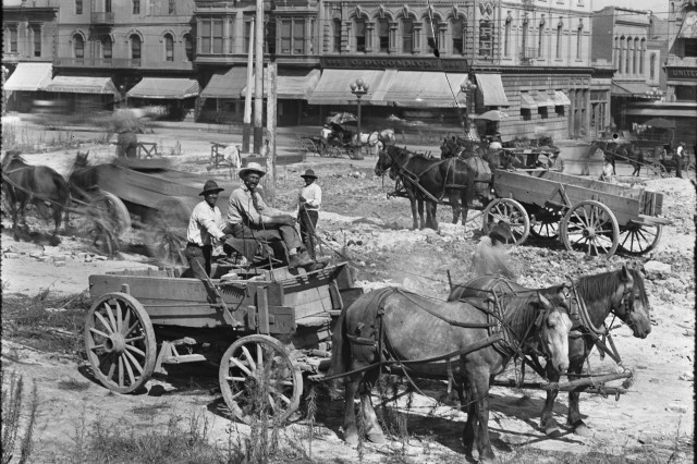 Antique photo of two men riding an open-top wooden wagon drawn by two horses across a rough dirt road. In the background are five other wagons crossing the road in front of a street corner with three-story buildings with awnings out front. 