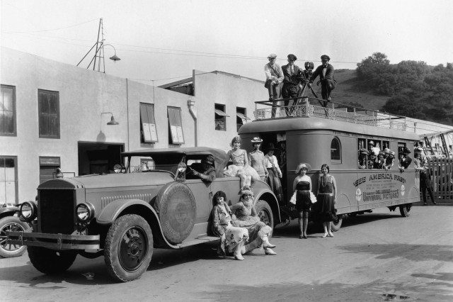 Automobile towing a trailer filled with actors; driver is at wheel and actresses sit on car; actresses and actors dressed in costume stand and look out of trailer; two men stand next to trailer while director and two crew members stand atop trailer with movie camera; building is to the left.