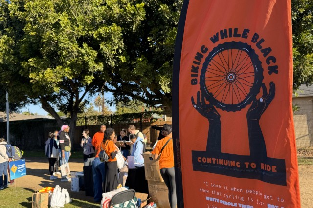 Orange feather banner in the foreground with the Biking While Black logo that are two black hands holding a spoked wheel. In the background are people milling about at a supply booth underneath large, leafy trees.