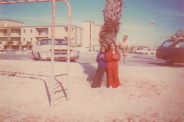 Two young girls, in jackets and long pants, one in blue and the other in red, standing in front of a palm tree at the beach, next to a set of metal monkey bars. Their father stands behind them tree, leaning into the photo next to his parked car.