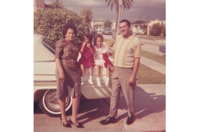 A family of color posing in their driveway in the early 1960s. The mother and father stand in front of a white sedan with their two preschool-age daughters in between them, sitting on the trunk of the car.