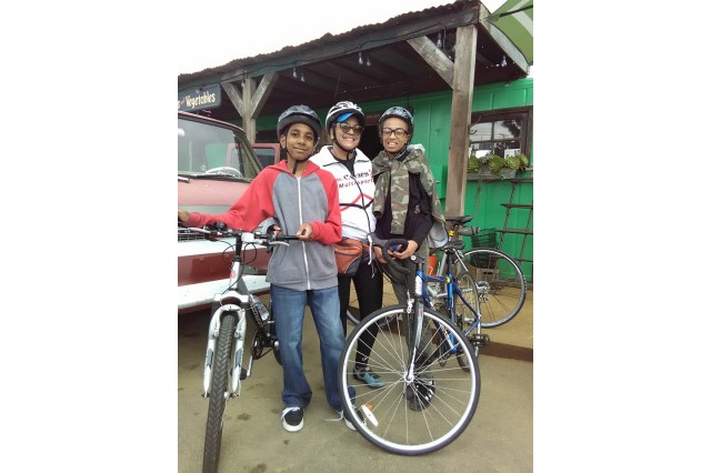 A woman and two kids of color standing next to their bikes in front of a a red car parked in front of a green building with a wooden awning.