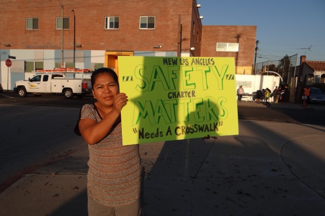 Woman of color standing in front of a brick school building, holding a yellow poster that reads &quot;Safety Matters: New Los Angeles Charter Needs a Crosswalk&quot;