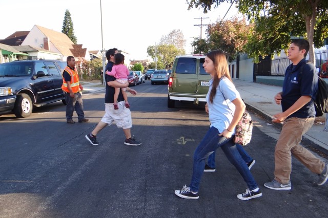 Parents and children walking across the street in front of a middle school with a crossing guard but no crosswalk