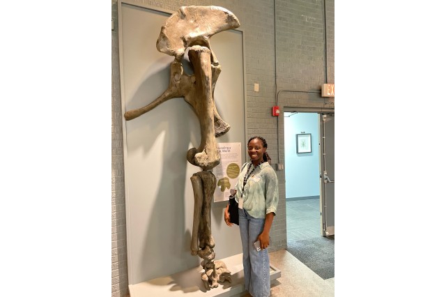 Intern in green button down shirt and jeans in front of bones mounted on the wall