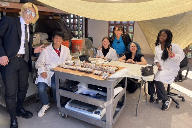 Interns in white lab coats around a table with specimens and two tv hosts