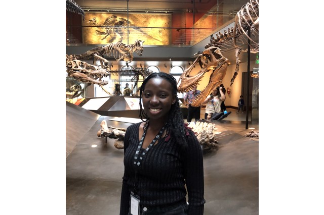 Ayomi Fadaka from the waist up, wearing a black long-sleeved shirt, with dinosaur skeletons in the background