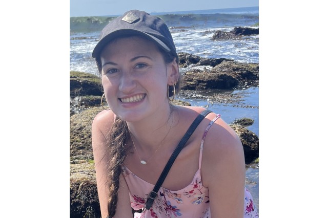 Head and shoulders portrait of Julia Morales wearing a navy baseball cap and pink tank top with rocky terrain and ocean in the background