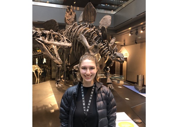 Libby Konjoyan from the waist up wearing a black shirt and puffy coat with dinosaur skeleton in the background