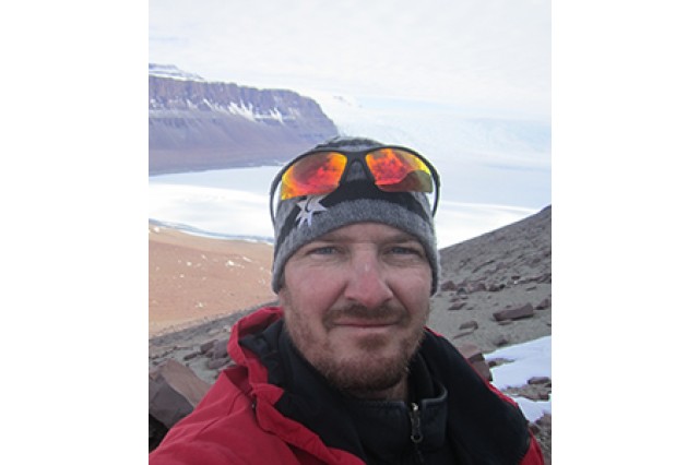 Nathan Smith wearing a gray beanie with orange tinted googles on his head and a red coat with the ocean and mountains in the background