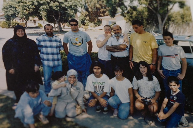 Multi-generational family photo with seven older members standing and seven younger members kneeling in front of them. The group poses on a suburban sidewalk in front of a blue and white station wagon.