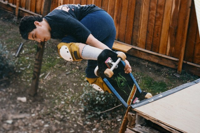 Teenage boy in dark t-shirt and shorts, with knee pads on, practicing skateboarding on a small ramp in his backyard.