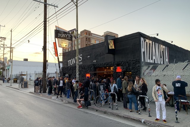 A line of customers wait outside a black brick restaurant at sunset with the name &quot;Pizzanista!&quot; in big white letters on the side of the building.