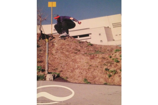 Skateboarder in a red and blue beanie, navy t-shirt, black jeans, and white shoes crouched over his skateboard high in the air with both feet on the board, his knees into his chest and his arms out to his sides. He wears a cast on his left arm. A small dirt hill is behind him, below a large, white square building. 