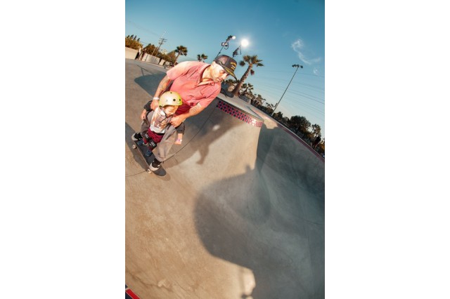 Father in a baseball hat, red shirt and khaki pants on a black skateboard, in a skate park, holding onto the shoulders of his young son who is also on the skateboard, in a yellow helmet, riding between his father&#039;s legs.
