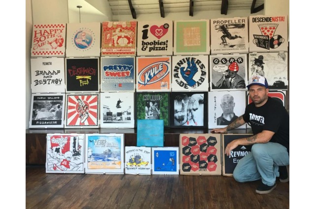 Man in baseball hat, black t-shirt, jeans &amp; black sneakers, kneeling next to a wall of 25 decorative pizza boxes