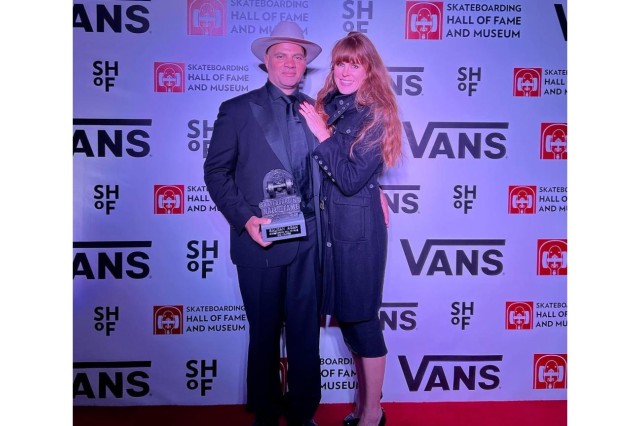 Red carpet photo of a man in a dark suit and white fedora hat, standing with his wife, who has long red hair and is wearing a long black coat and black capri pants. He is holding a small trophy shaped like half of a skateboard that reads &quot;Skateboarding Hall of Fame&quot; in front of a white backdrop with the Vans company logo on it.