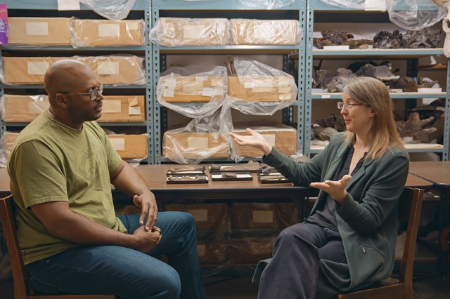 Shane Campbell-Staton sitting at a table, facing Emily Lindsey, with shelves of specimens in the background