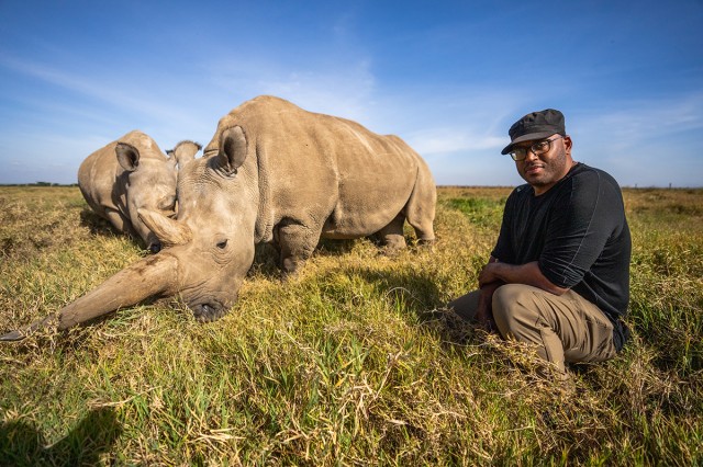 Shane Campbell-Staton in a field knelling next to two rhinos with blue sky behind them
