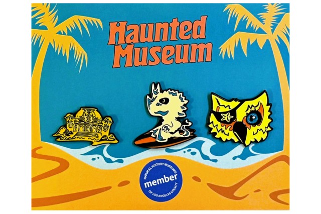 Haunted Museum in orange letters above a mansion, ghost on a surfboard, and owl pins with yellow palm trees on either side against a blue, ocean, and sand background